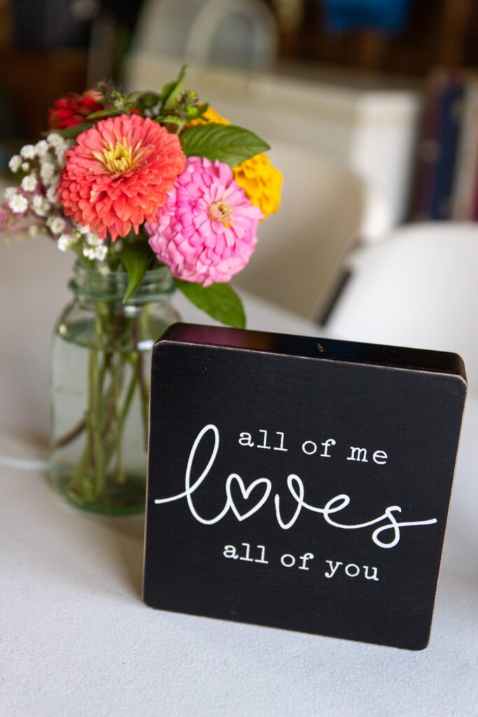 "all of me loves all of you" sign next to a jar of colorful zinnias at backyard wedding